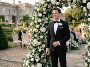 Handsome groom in classic black tuxedo standing near floral wedding arch