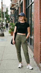 Young woman in fitted crop top, joggers, and baseball cap, relaxed pose, urban street background, athleisure style, soft natural lighting